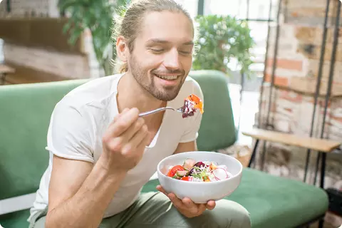 Jeune homme assis sur le canapé, riant et mangeant une salade colorée