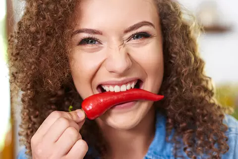 Frau mit braunen Locken hat eine Chili zwischen den Zähnen und lacht