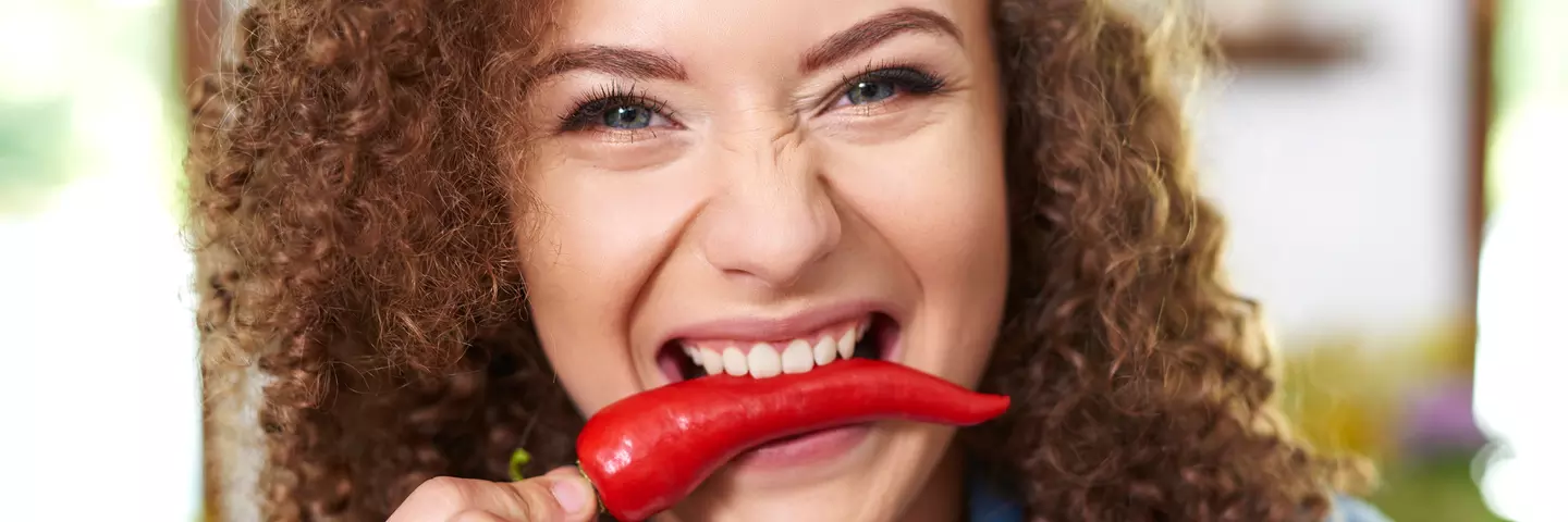 Frau mit braunen Locken hat eine Chili zwischen den Zähnen und lacht