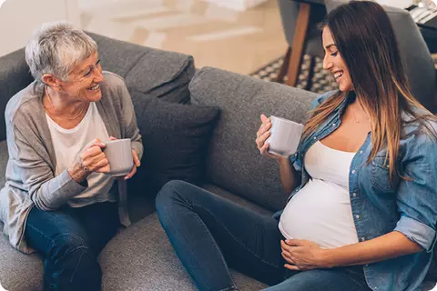 Eine ältere Frau und eine Schwangere sitzen lachend auf dem Sofa und trinken Tee