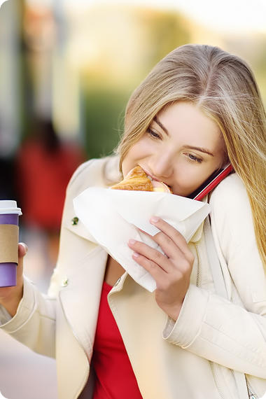 Une jeune femme avec un téléphone portable à l'oreille mange un hamburger à la hâte - Les hormones et la nourriture hâtive peuvent provoquer des brûlures d'estomac