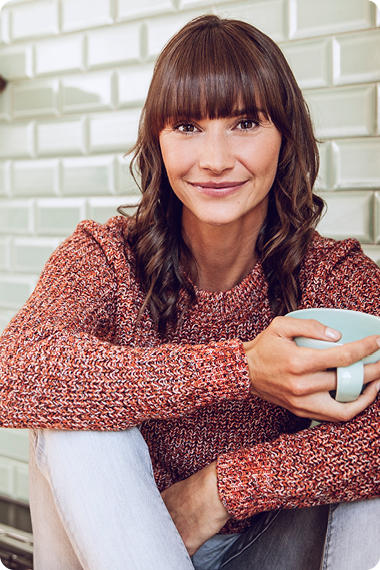 Frau mit langen braunen Haaren und Pullover sitzt entspannt mit Kaffeetasse auf dem Boden - wie gestalte ich meinen Alltag